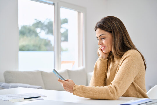 Happy middle aged woman using mobile cell phone sitting at table in living room. Smiling mature lady holding smartphone buying online doing shopping in smartphone app, texting in social media at home.