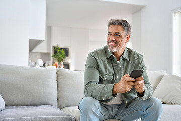 Happy older mature middle aged man holding cell phone using smartphone sitting at home on sofa, relaxing on couch in living room, looking away at copy space advertising mobile offers and apps.