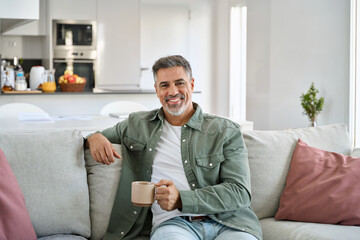 Happy middle aged man holding coffee cup relaxing on couch at home. Smiling mature older man...