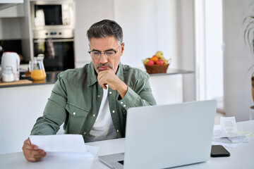 Worried stressed middle aged mature man wearing eyeglasses looking at paper bill checking insurance...