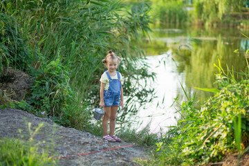 Fototapeta na wymiar Three-year-old blond girl in denim shorts with straps catches fish on the river 