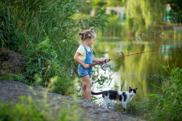 Three-year-old blond girl in denim shorts with straps catches fish on the river with a black and...