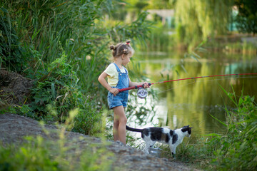 Three-year-old blond girl in denim shorts with straps catches fish on the river with a black and...