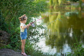 Fototapeta na wymiar Three-year-old blond girl in denim shorts with straps catches fish on the river with a black and white cat