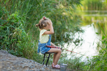 Three-year-old blond girl in denim shorts with straps catches fish on the river 