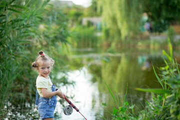 Three-year-old blond girl in denim shorts with straps catches fish on the river 