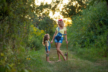 Two blond barefoot fisherwomen in denim shorts with straps with a fishing rod and bucket on a road surrounded by greenery against the backdrop of the setting sun