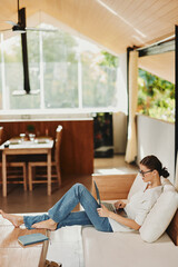 Woman using laptop on a cozy couch in her modern home office She is a successful freelancer working on her startup with a smile, surrounded by technology and wireless devices The room has a nice