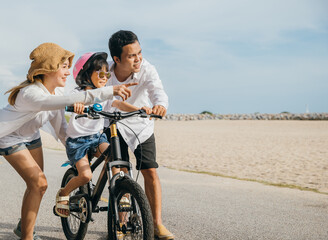 Obraz premium On sandy beach near sea cheerful family enjoys summer road trip teaching and learning bicycle riding. Parents guide their children creating joyful and memorable vacation moment.