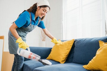 In modern setting Asian woman maid uses a vacuum machine cleaner to clean a sofa in a living room. Her dedication to hygiene and housework shines through as she focuses on furniture care.