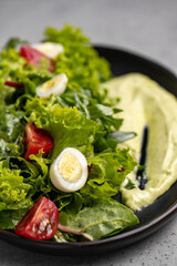Fresh green salad with quail eggs, cherry tomatoes and guacamole in black plate on grey background