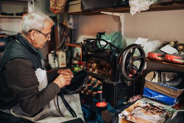 An old artisan is sitting at cobbler's workshop and repairing a purse.