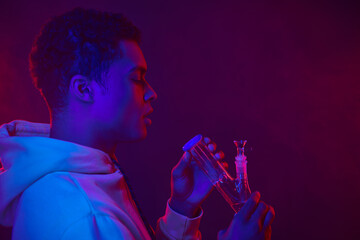 young african american man in hoodie looking at glass bong on dark purple background with lighting