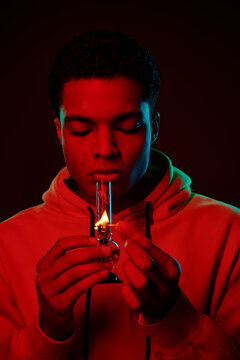 young african american man in hoodie lighting glass bong on dark background with red lighting
