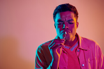 focused african american guy lighting cigarette with match under blue neon lights on pink background