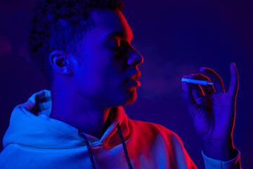 african american man holding rolled cigarette while standing on dark blue background with neon light - Powered by Adobe