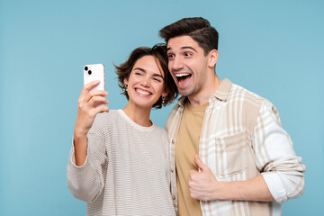 Young smiling friends hugging holding mobile phone making selfie over blue wall background - obrazy, fototapety, plakaty