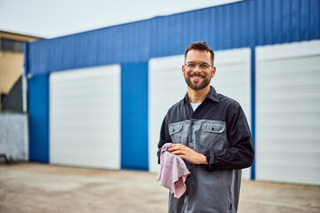 Portrait of a happy mechanic man, standing in front of his car repair shop, holding a cloth.