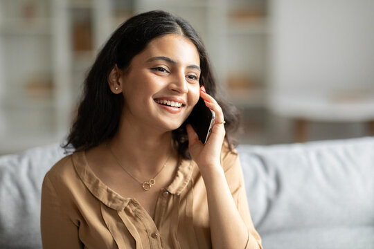Smiling indian woman talking on cellphone, enjoying pleasant mobile conversation while resting on couch at home in living room, copy space