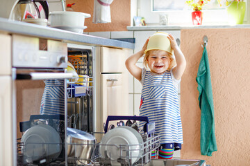 Little adorable cute toddler girl helping to unload dishwasher. Funny happy child standing in the kitchen, holding dishes and putting a bowl on head. Healthy kid at home. Gorgeous helper having fun - Powered by Adobe