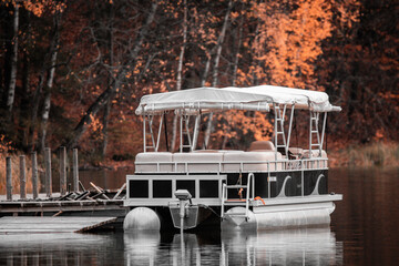 The boat anchored by the pontoon floats on the lake against the background of trees with colorful leaves

