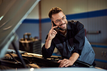 A smiling mechanic looking on the side, making a phone call while working at his garage, checking a car.
