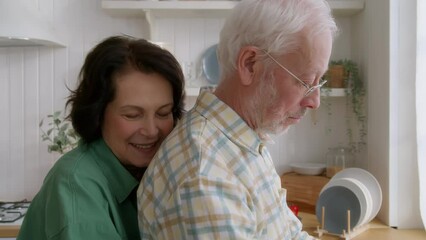 elderly couple spend time together in kitchen during morning routine. old man wash fruits, his wife comes up to embrace his back. long life love relationship, tender moments