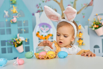 Fototapeta na wymiar Baby girl celebrate Easter. Funny happy kid in bunny ears Sits by the table and playing with Easter eggs. Colorful Easter eggs and flowers. Home decoration and flowers