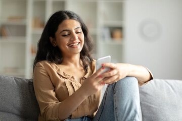 Smiling hindu lady using her smartphone while relaxing on couch at home, happy woman looking online...