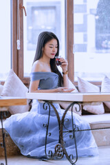 Beautiful princess in blue dress sitting and drinking a hot beverage while looking out of the window.Young Asian woman sipping hot tea at a table by the window.Relaxing time sipping hot tea or coffee.