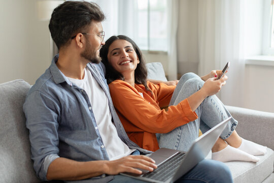 Weekend pastimes. Happy indian spouses using laptop and smartphone at home, resting on sofa and looking at each other, spouses browsing internet
