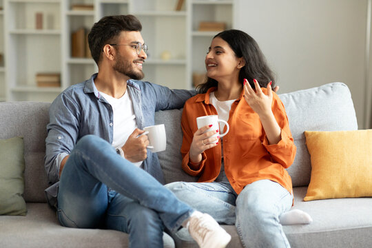 Loving hindu spouses drinking coffee and chatting while sitting on sofa at home, enjoying spending time together on weekend. Happy romantic relationship