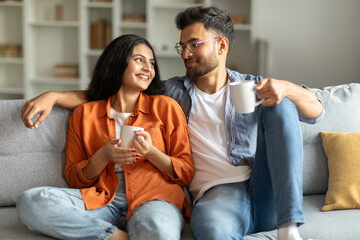 Weekend morning. Happy hindu spouses enjoying hot coffee and talking, sitting on sofa, holding cups...