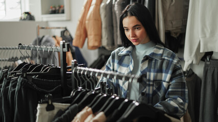 Beautiful brunette girl stands in a fashion store, choosing clothing items to buy. She browses...