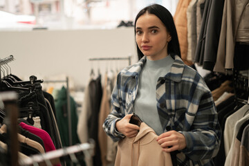 A young woman makes purchases, choosing clothes at a discount in a clothing store, stands looking at the camera, thinking