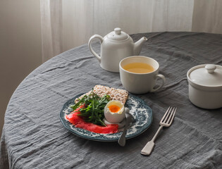 Delicious homemade breakfast, brunch - boiled egg, lightly salted salmon, arugula, bread rolls and green tea on a round table with a gray muslin tablecloth
