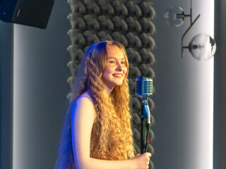 A young girl with blonde curly hair sings into a retro microphone. Performance of a girl with long hair on stage. The performance of the song at the concert. A smile on the girl's face.