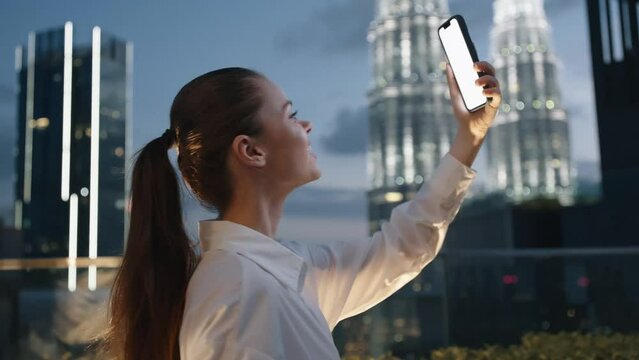 A woman happy stares into a phone screen in her hands shooting a video on her phone on the roof of a skyscraper overlooking the big city and the glowing buildings, the lifestyle of the night city.