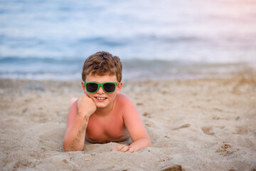 Little boy kid with sunglasses is lying on the sand near seaside smiling after sunburn.