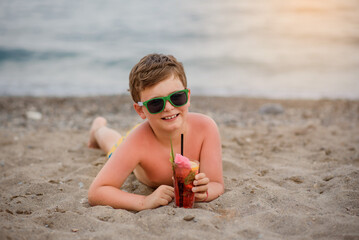 Little boy kid with sunglasses is lying on the sand near seaside smiling and drinking a cocktail.