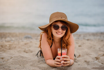 Young beautiful woman wearing sunglasses and a hat is lying on beach sand and drinking a cocktail near a tropical sea.