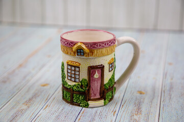A ceramic mug with a handle with a three-dimensional image of the windows of a wooden house with a fence on a gray wooden table. Design, painting, applied art, ceramics.