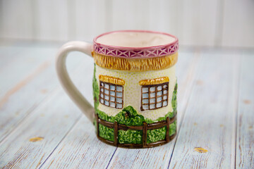 A ceramic mug with a handle with a three-dimensional image of the windows of a wooden house with a fence on a gray wooden table. Design, painting, applied art, ceramics.