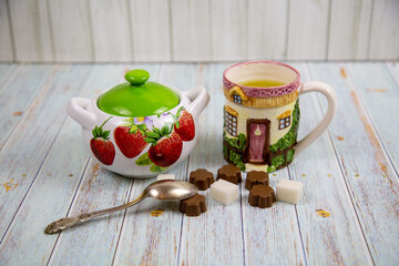A ceramic mug with a handle depicting the windows of a wooden house, a sugar bowl with strawberries, sugar and sweets on a gray wooden table. Design, painting, applied art, ceramics.