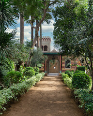A pathway winds through lush greenery, leading to a Mamluk style building nestled among trees and bushes, creating a serene landscape with a mix of plants and fixtures