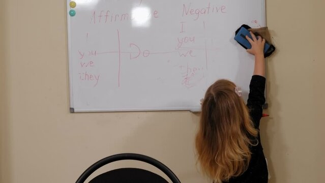 A cute little girl wipes the board with a washcloth in elementary school, a rule is written on the white board with a marker. A lesson in elementary school.