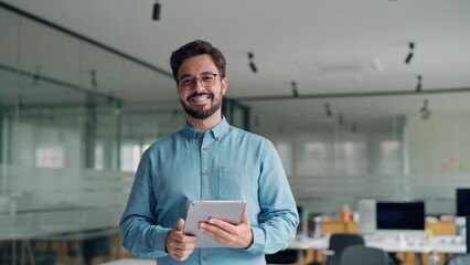 Smiling busy young latin business man manager using tablet computer, happy hispanic businessman executive looking at camera holding tab working standing in office. Portrait. - Powered by Adobe