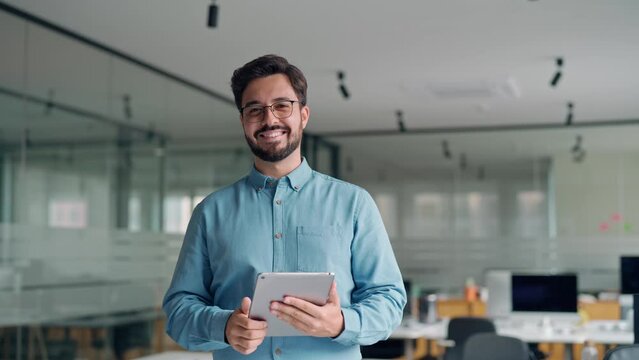 Smiling busy young latin business man manager using tablet computer, happy hispanic businessman executive looking at camera holding tab working standing in office. Portrait.