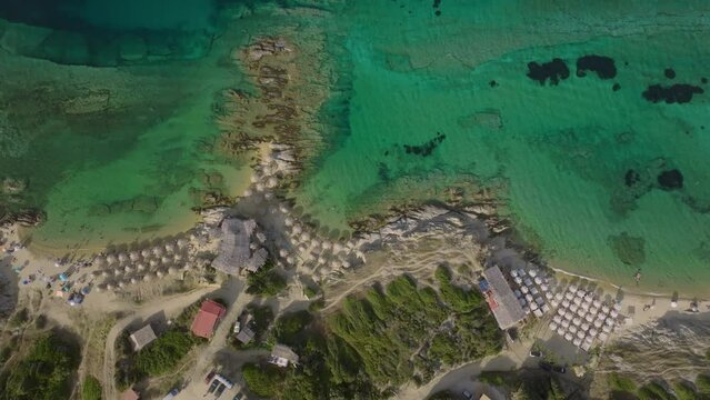 An aerial shot from an ascending drone presents the tranquil beachfront, where clear turquoise waters kiss the soft sandy shores. Rows of umbrellas and beachgoers enjoying the serene environment