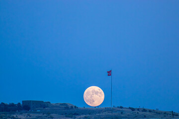 This captivating image captures the full moon rising majestically behind a Turkish flag atop an...